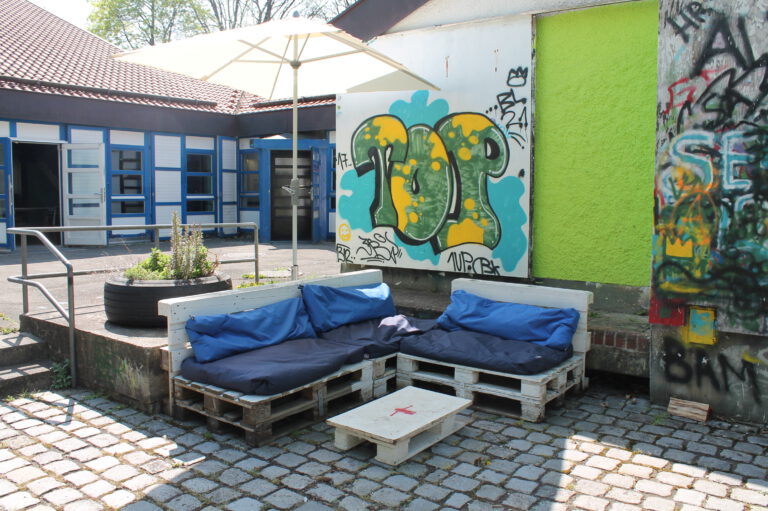 sommerliches Foto vom Hof des Treff21 mit Palletten-Couch, Graffitiwand und offenen Türen