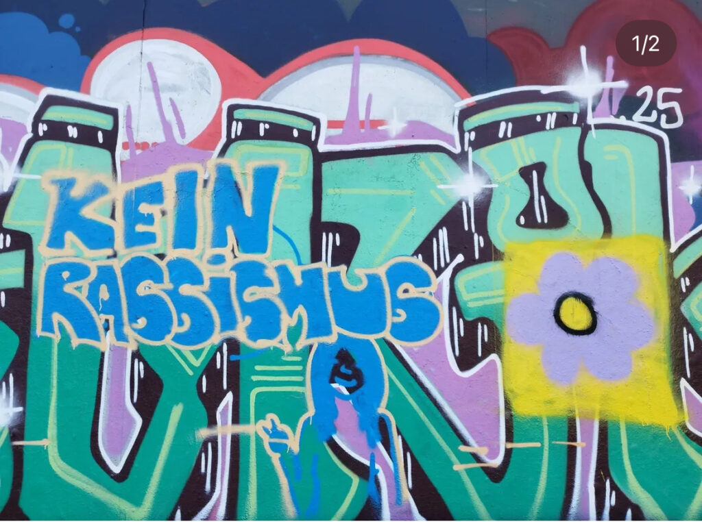 Plakat zur Ausstellung "Doppelt unsichtbar" im Hintergrund ein buntes Graffitibild mit dem Schriftzug "Kein Rassismus"